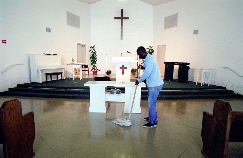 A Black person with a shaved head wearing a blue button down shirt, jeans, and black sneakers mops the floor of a church in front of the pulpit. A large hanging cross on the wall, podium, and white piano are on the stage behind them.