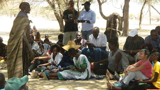 Community members sit on the ground, listening to a speaker wrapped in a striped cloth, The speaker is Black and has a bald head.