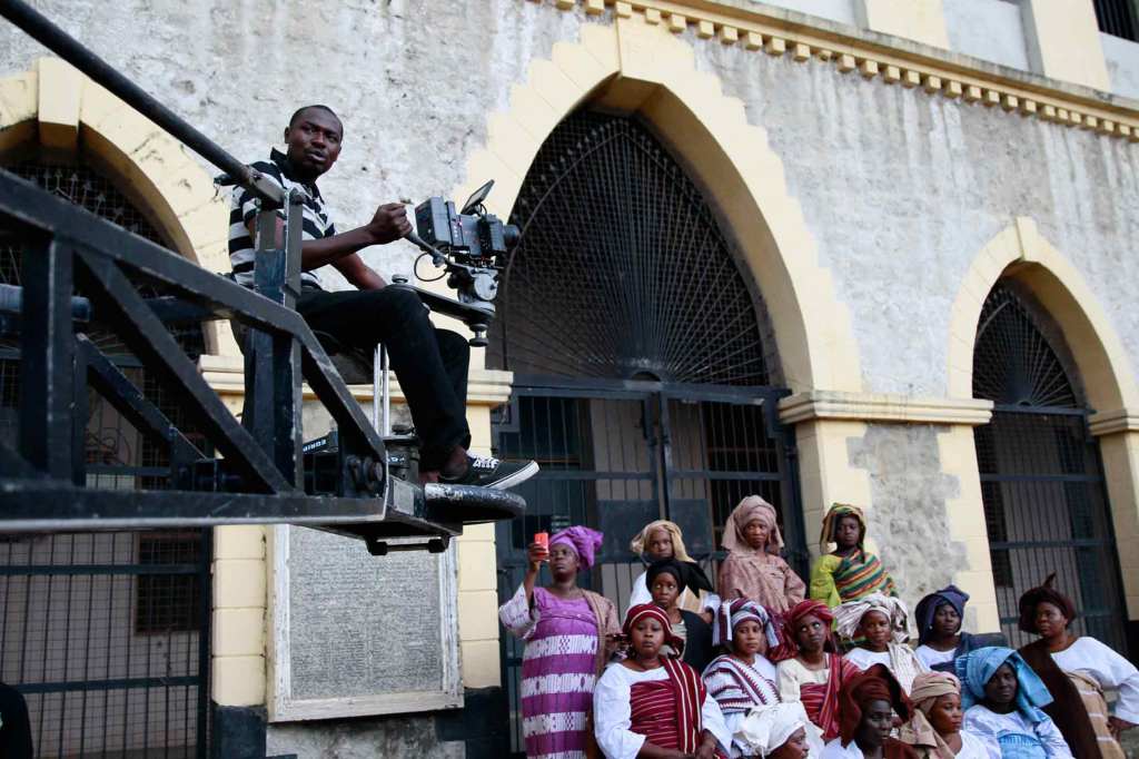 A Black cameraperson in a striped polo, dark jeans, and sneakers films a scene from a crane during the making of "Ake" in Nigeria. Multiple Black people wearing colorful traditional Nigerian women's clothing sit and watch.