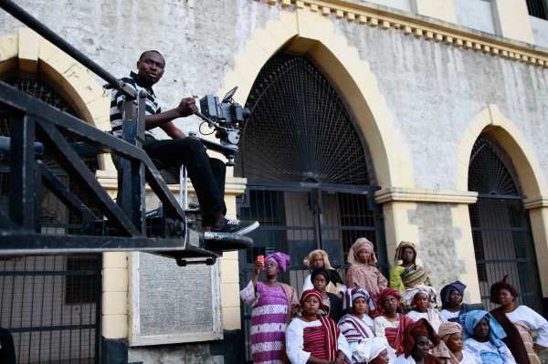 A Black cameraperson in a striped polo, dark jeans, and sneakers films a scene from a crane during the making of "Ake" in Nigeria. Multiple Black people wearing colorful traditional Nigerian women's clothing sit and watch. 