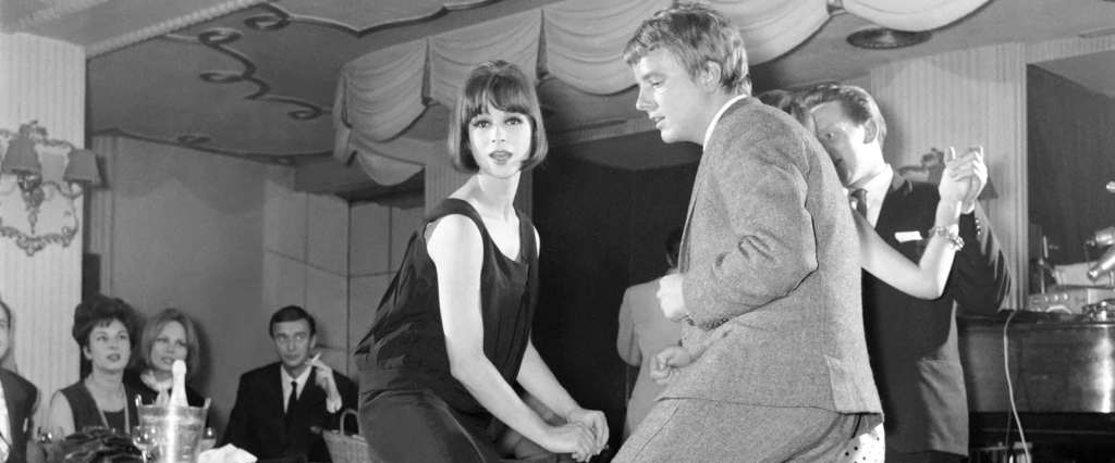 A black and white picture of a young couple doing the twist. One is wearing a loose tank dress and the other is in a light colored suit.