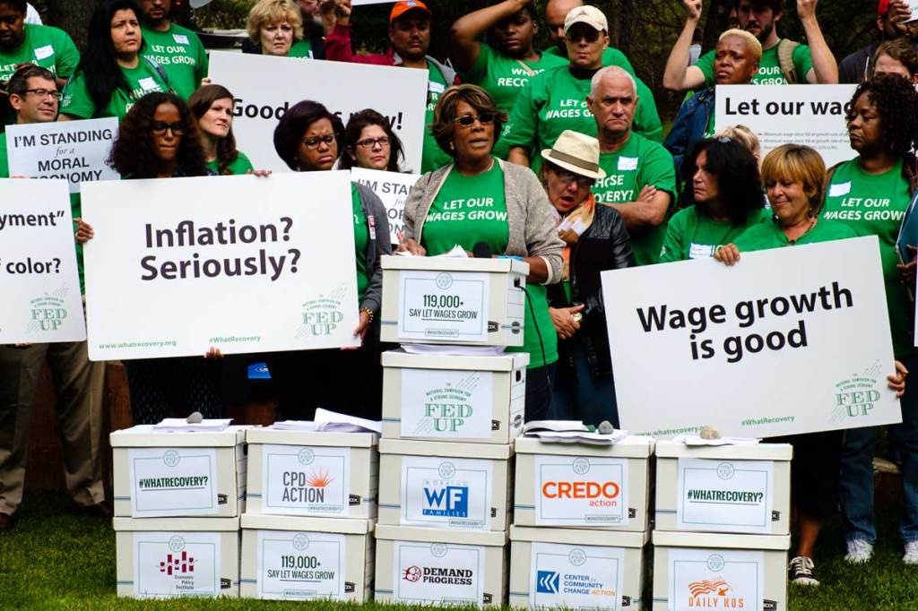 Protesters in green t-shirts that read "LET OUR WAGES GROW"  hold printed Fed Up campaign signs stand behind a speaker, a Black person with a short dark bob, the blazer and the same green t-shirt. They stand at a podium made of office filing boxes.