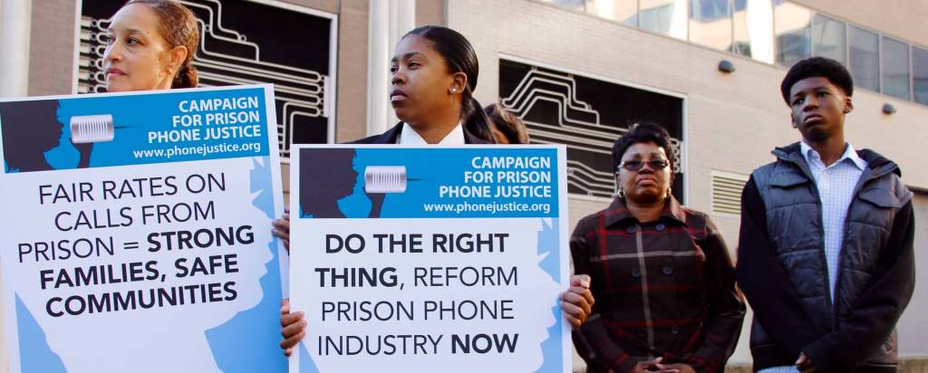 Four Black protesters stand with a somber look on their faces. Two demonstraters hold signs that read "Campaign for Prison Phone Justice, www.phonejustice.org, Fair rates on calls from prison = strong families, safe communities" and "Do the right thing, reform prison phone industry now."