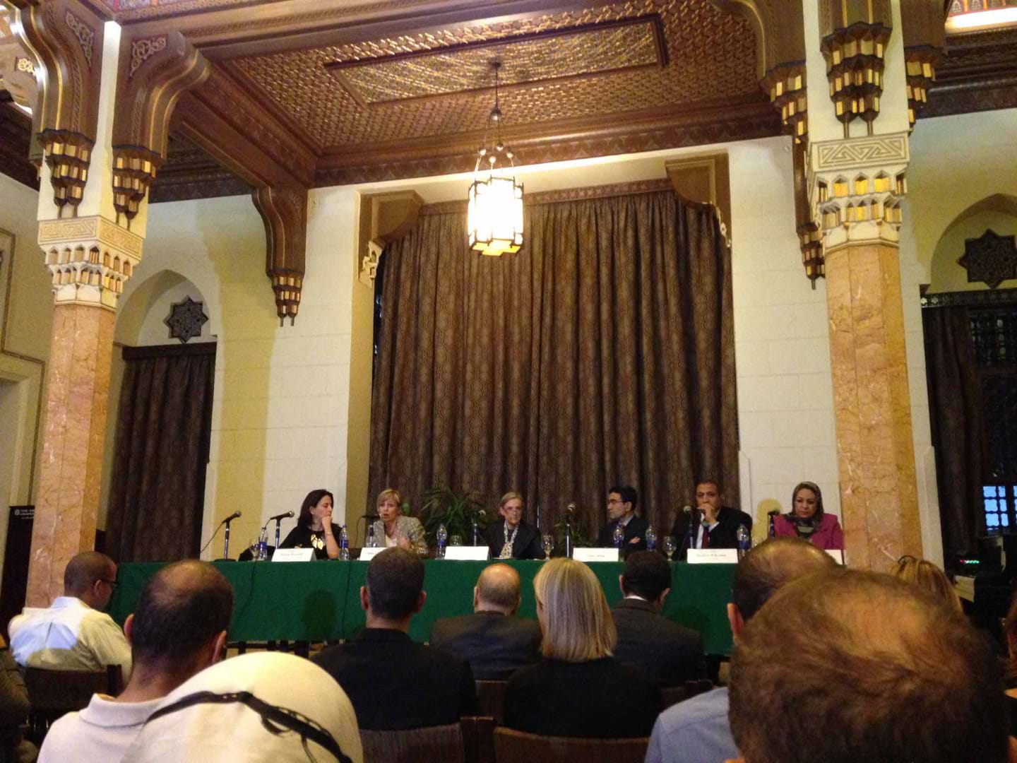 A panel of speakers sits behind a long table with dark green tablecloth. They each have a microphone and name tag on the table in front of them.