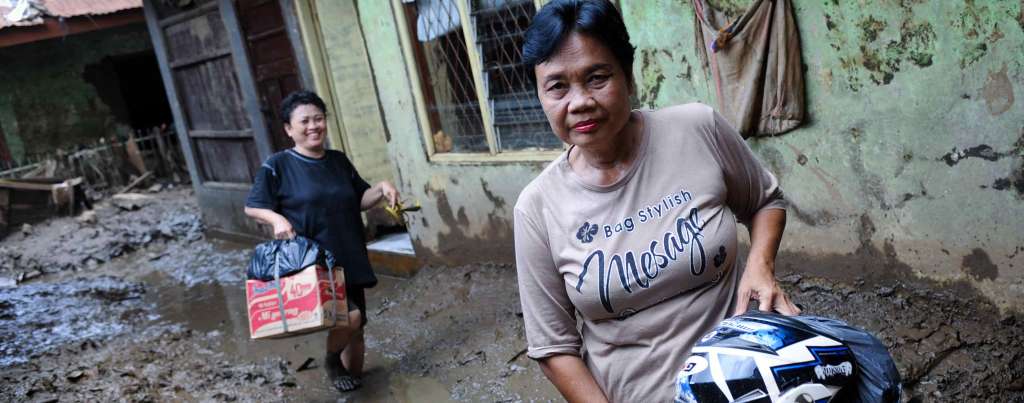 Two Southeast Asian people hold their belongings while walking through a street in an Indonesian flood zone. The ground is covered in wet mud.