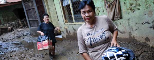 Two Southeast Asian people hold their belongings while walking through a street in an Indonesian flood zone. The ground is covered in wet mud. 