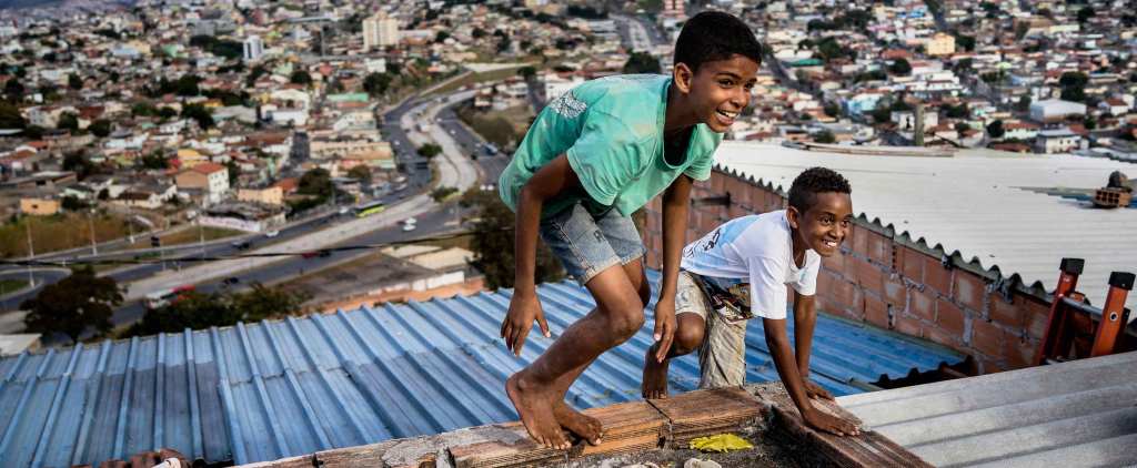 Two smiling children with medium dark skin tones and short hair play on the rooftop of a favela. They are barefoot, and wear t-shirts and shorts.