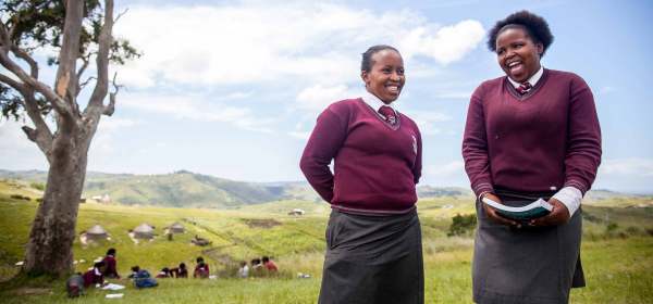 Two Black students stand smiling in a grassy field in their school uniforms, grey skirts, white collared shirts, and maroon sweaters with ties. One carries a paperback textbook in their hands.