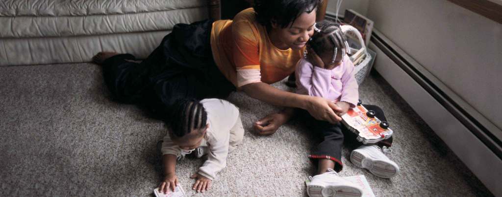 A Black parent and their young Black children play on the floor. One child with beads in their braids and a lilac colored shirt holds a book shaped like a bus with wheels on it. The other child with cornrows crawls on the gray carpet.