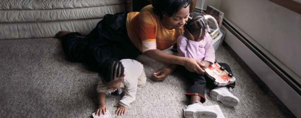 A Black parent and their young Black children play on the floor. One child with beads in their braids and a lilac colored shirt holds a book shaped like a bus with wheels on it. The other child with cornrows crawls on the gray carpet. 