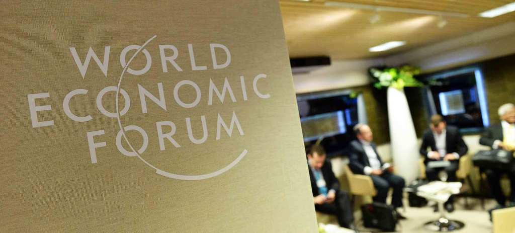 The World Economic Forum logo is on a sign. Out of focus in the background, four people in suits sit in chairs.