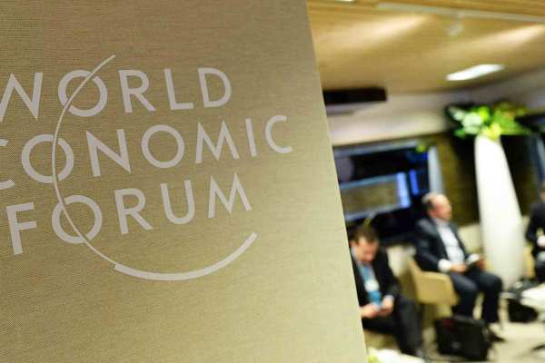 The World Economic Forum logo is on a sign. Out of focus in the background, four people in suits sit in chairs. 
