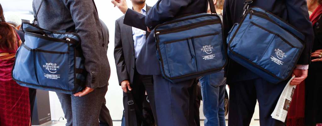 A close up of blue messenger bags with th World Economic Forum logo printed on them being carried by three people in suits.