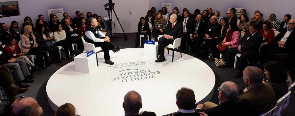 Shimon Peres sits on a circular white stage facing an interviewer at the World Economic Forum. The stage is surrounded by a seated audience.