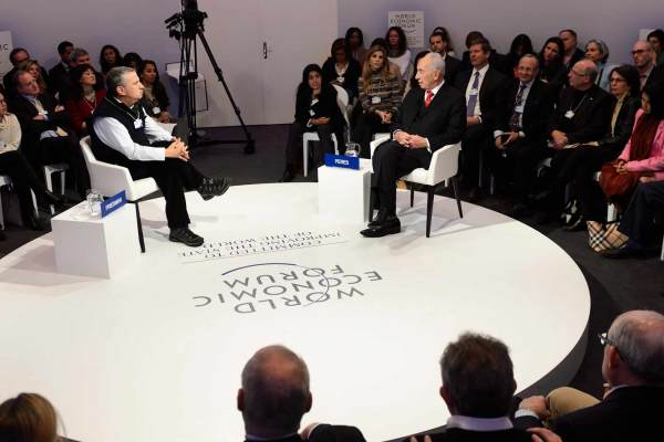 Shimon Peres sits on a circular white stage facing an interviewer at the World Economic Forum. The stage is surrounded by a seated audience. 