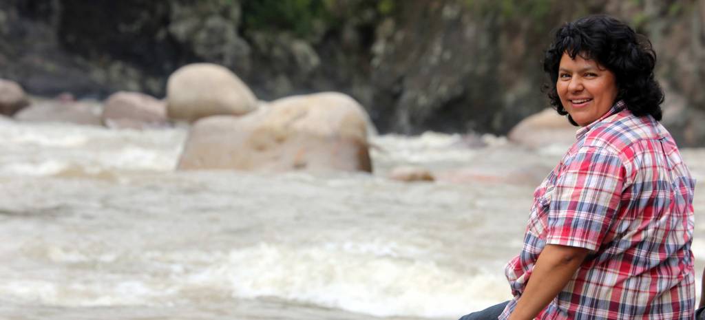 Berta Cáceres sits on the banks of Gualcarque River in western Honduras. She faces the river and turns her smiling face towards the camera. She wear a red, white, and blue plaid short sleeved shirt.