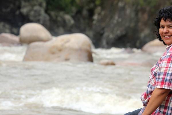 Berta Cáceres sits on the banks of Gualcarque River in western Honduras. She faces the river and turns her smiling face towards the camera. She wear a red, white, and blue plaid short sleeved shirt.
