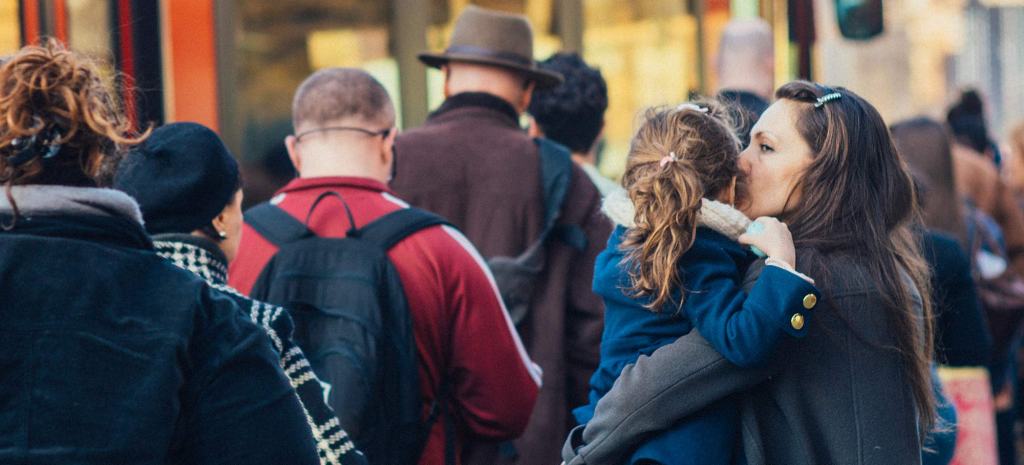 A crowd of people stands in a queue with their backs to the camera. An adult with a blue barrette in their long brown hair carries a child in a royal blue coat with their light brown hair in a ponytail.