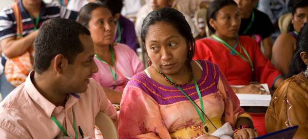 Indigenous activists in Peru sit at an oudoor conference wearing green lanyards. Two people sit in the foreground, one in a pink polo and short hair, another with their long hair tied back in a pink and yellow dress trimmed with maroon and orange on the neckline. 