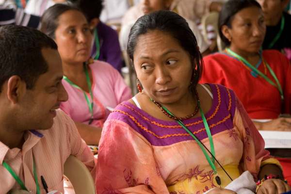 Indigenous activists in Peru sit at an oudoor conference wearing green lanyards. Two people sit in the foreground, one in a pink polo and short hair, another with their long hair tied back in a pink and yellow dress trimmed with maroon and orange on the neckline. 