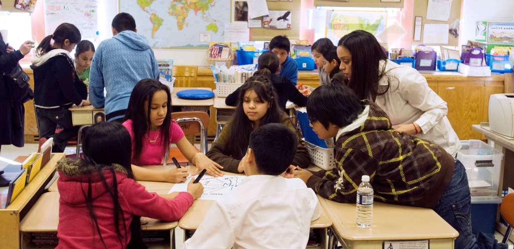 Young Black and brown students gather around a cluster of desks in a classroom. An adult with long straight black hair leans over the table to assist them.