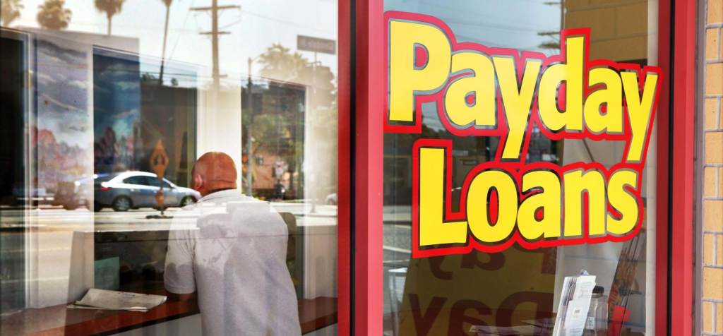 The front window of a storefront reads "Payday Loans" in bright yellow letters with a red outline.  Behind the glass, a bald person in a white t-shirt stands at the reception desk with their back to the window.