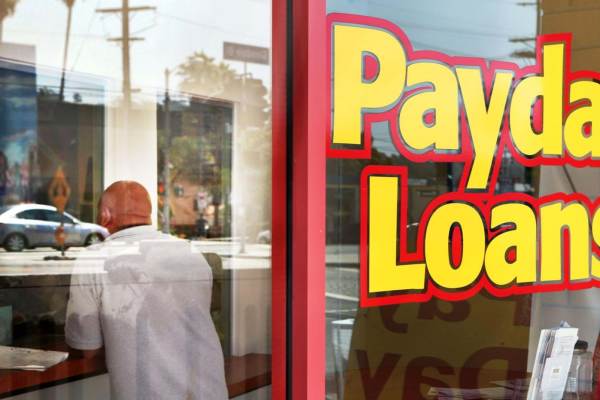 The front window of a storefront reads "Payday Loans" in bright yellow letters with a red outline.  Behind the glass, a bald person in a white t-shirt stands at the reception desk with their back to the window. 