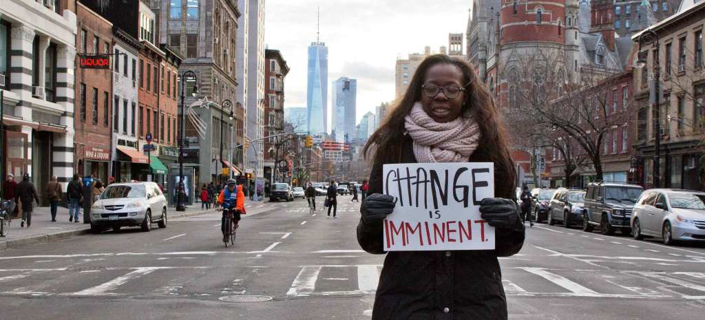 A Black person with long straight hair stands in a city crosswalk holding a handmade sign that reads "Change is Imminent."