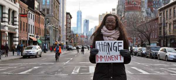 A Black person with long straight hair stands in a city crosswalk holding a handmade sign that reads "Change is Imminent." 