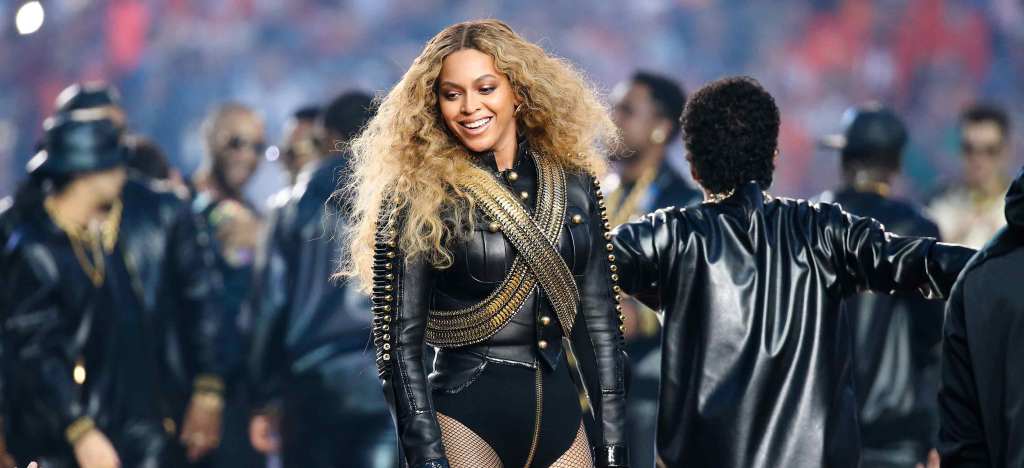 Beyonce stands smiling onstage at the Super Bowl Halftime show. She is wearing a black leather jacket with gold accents.