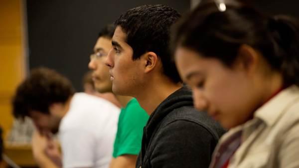 A person with medium skin tone and black hair sits in a classroom with other students listening to a lecture. 