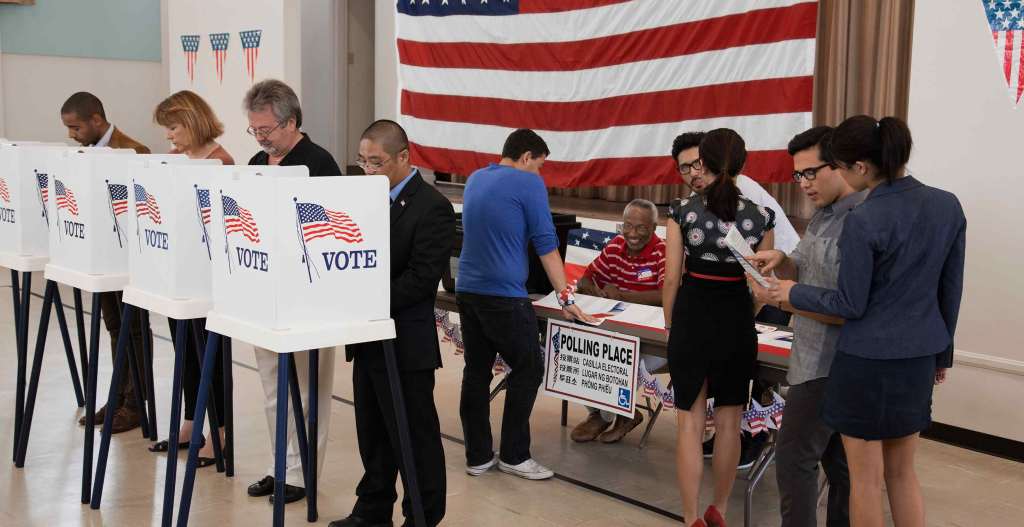 Voters gather at United States polling place. Four people stand at individual voting booths decorated with U.S. flags and the word VOTE. A smiling polling place worker sits at a folding table in front of a large U.S. flag on the wall. A line of voters stands in front of the table.
