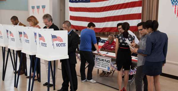 Voters gather at United States polling place. Four people stand at individual voting booths decorated with U.S. flags and the word VOTE. A smiling polling place worker sits at a folding table in front of a large U.S. flag on the wall. A line of voters stands in front of the table. 