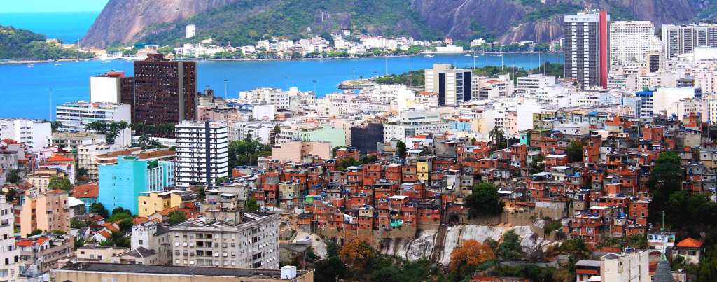 A sweeping view of downtown Rio De Janeiro and the Sugarloaf/Pain de Sucre Favéla in Brasil. The colorful buildings are flanked by deep blue sea in the harbor, with mountains and white buildings across the water.