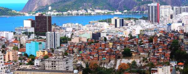 A sweeping view of downtown Rio De Janeiro and the Sugarloaf/Pain de Sucre Favéla in Brasil. The colorful buildings are flanked by deep blue sea in the harbor, with mountains and white buildings across the water. 