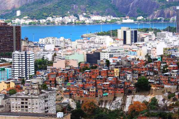 A sweeping view of downtown Rio De Janeiro and the Sugarloaf/Pain de Sucre Favéla in Brasil. The colorful buildings are flanked by deep blue sea in the harbor, with mountains and white buildings across the water. 