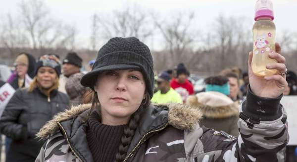 A Flint, Michigan protester wearing winter clothes holds a baby bottle full of dirty water. They have their hair in a long dark braid and a facial piercing beneath their left eye. 