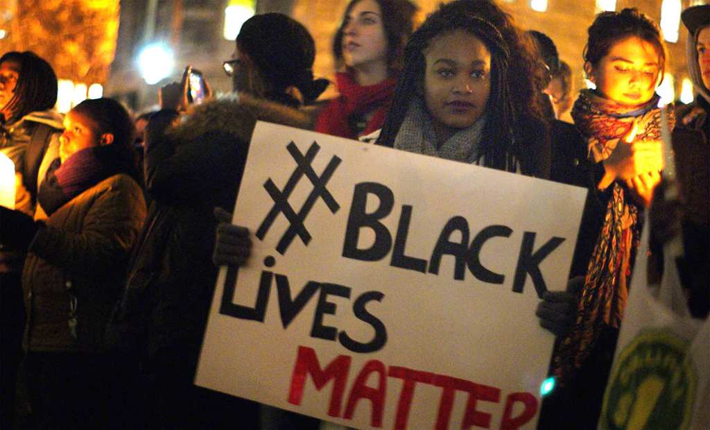 A Black person with box braids and gloves holds up a homemade sign that reads #BlackLIvesMatter at a candlelight vigil with other people paying their respects.