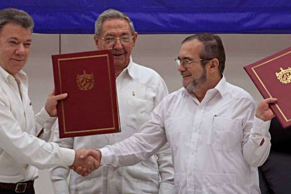 Juan Manuel Santos, Raúl Castro, and Timoleón Jimenez smile on stage and shake hands while holding red portfolios with gold trim.