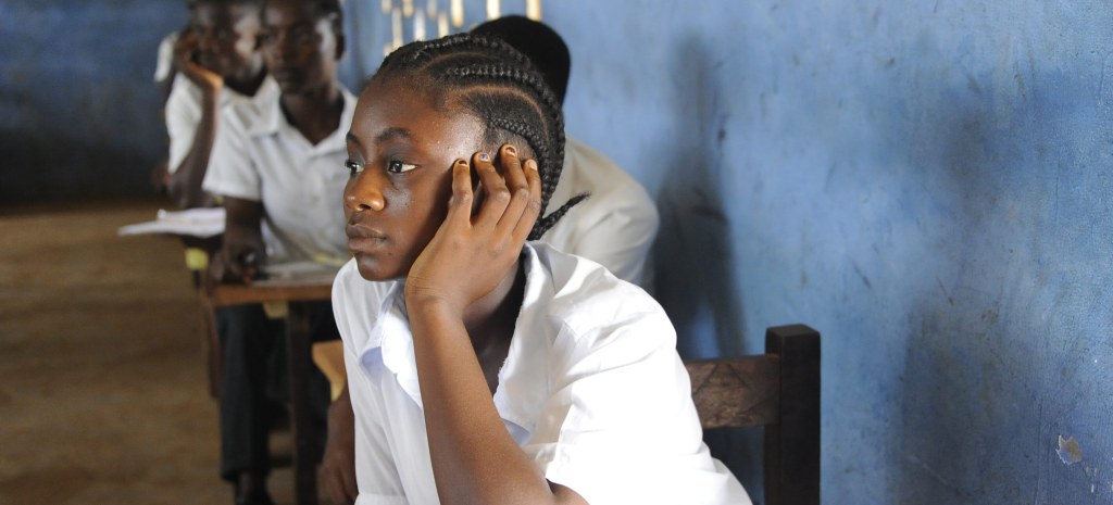A Black student in a white short sleeved polo shirt and cornrows, pays attention at their desk in a classroom. They rest their head on their hand as they focus intently.