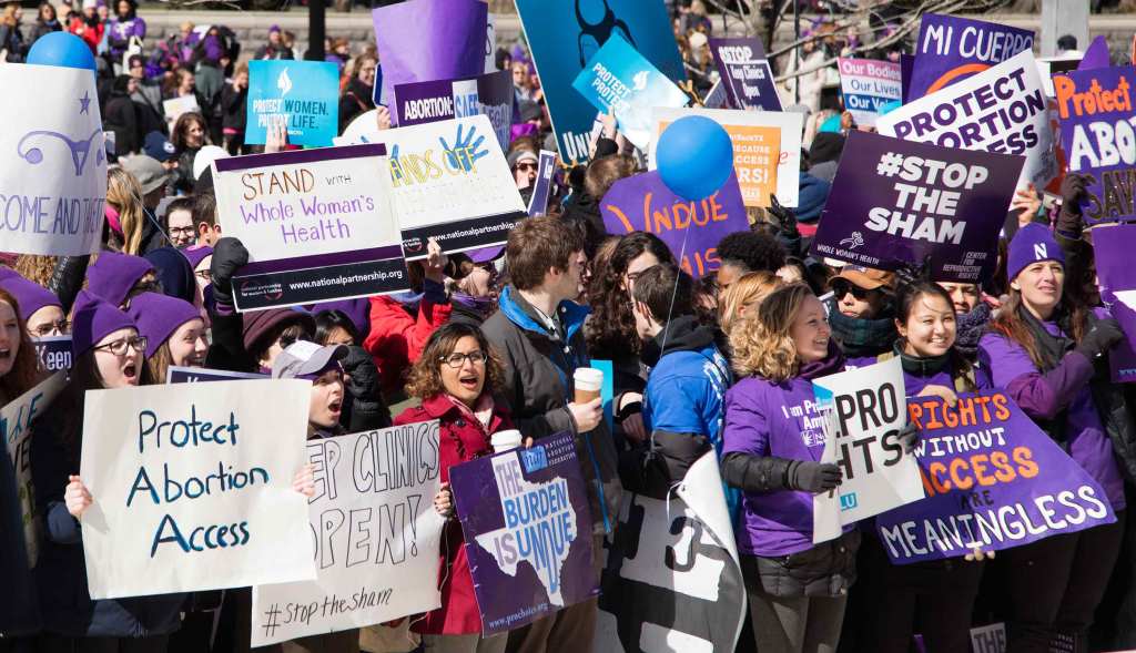 Demonstrators in winter clothes stand outside of the U.S. Supreme Court. They hold a mix of handmade and professionally made signs in support of reproductive rights and abortion access, including signs that read "Protect Abortion Access", "Keep Clinics Open! #StopTheSham", "The Burden is Undue" in the silhouette of the state of Texas, "Rights without Access are Meaningless" and "Stand with Whole Woman's Health".