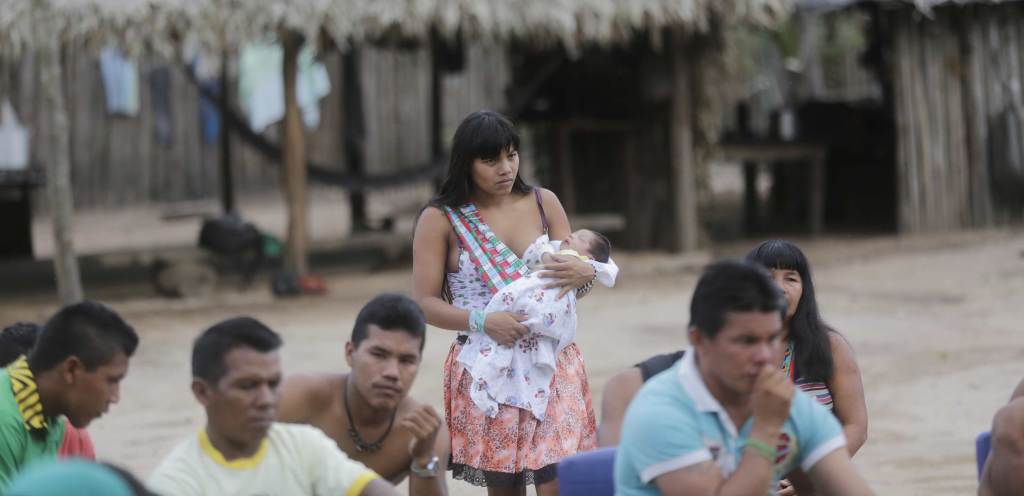 Members of the indigenous Ka'apor community in Brazil’s Alto Turiaçu reserve gather outside to watch something. Most are sitting, but one person wearing a white shirt and patterned orange skirt stands, holding an infant wrapped in a printed white blanket.