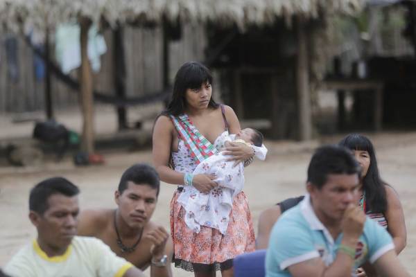 Members of the indigenous Ka'apor community in Brazil’s Alto Turiaçu reserve gather outside to watch something. Most are sitting, but one person wearing a white shirt and patterned orange skirt stands, holding an infant wrapped in a printed white blanket. 