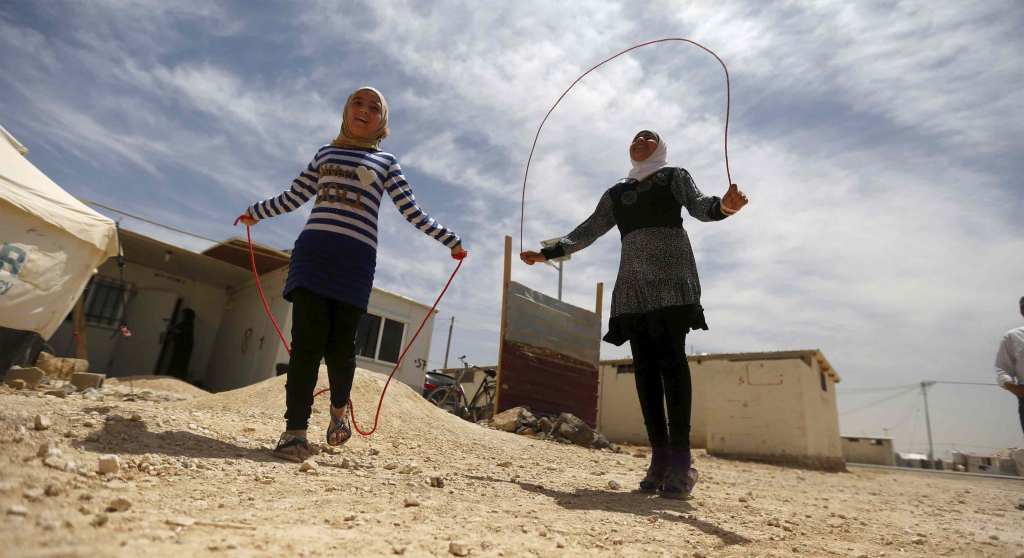 Omayma al Hushan, a teenage Syrian girl, plays outside with another girl. Both girls are wearing hijabs and jumping rope beneath a blue sky streaked with clouds.