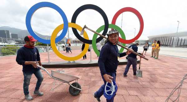 Two workers in jeans and hooded sweatshirts walk past a large sculpture of the Olympic rings logo installed in a brick courtyard. One carries a hoe slung over their shoulder in one hand and a hard hat in the other. The other pushes a metal wheelbarrow.