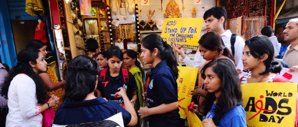 Indian youth gather in a Bangalore street holding bright yellow signs that read "World AIDS Day" and "AIDS: Stand up for the Challenge. It's Everyone's Responsibility." They stand outside of a store window with brightly colored jewelry on display.