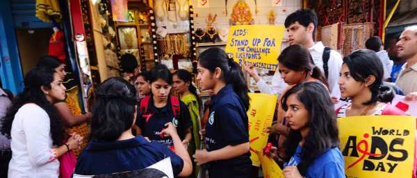 Indian youth gather in a Bangalore street holding bright yellow signs that read "World AIDS Day" and "AIDS: Stand up for the Challenge. It's Everyone's Responsibility." They stand outside of a store window with brightly colored jewelry on display.