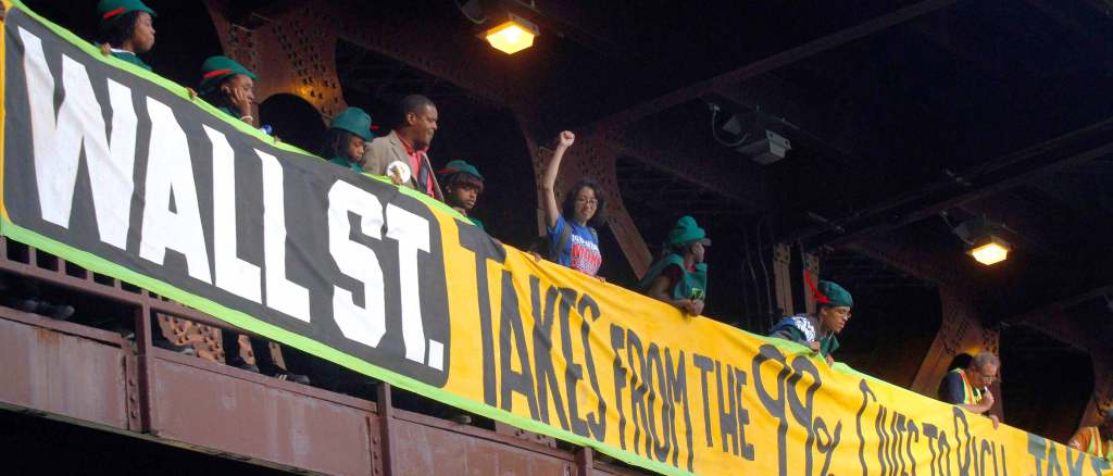 Protestors stand behind a banner that reads "Wall St. Takes from the 99%" hung from the side of a bridge. Black children and teens wear green Robin Hood hats with red feathers. One protestor with short dark hair and glasses looks below and raises a fist.