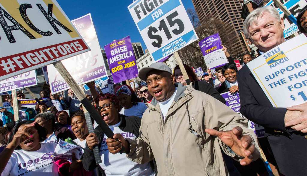 A packed crowd of protesters march in the street, smiling and yelling. Many hold signs supporting fair wages, including signs that read "#FightFor15", "Support the Fight for $15 , SEIU NAGE" and "32BJ SEIU, Justice for Janitors, Raise America with Good Jobs"