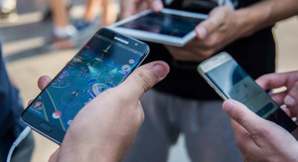 A close up shot of hands holding mobile devices with Pokemon Go on the screen. Three people stand in a circle, two hold smartphones and a third holds a tablet. 